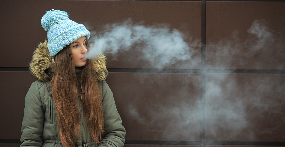 Teen vaping. Young girl smoking an electronic cigarette opposite modern brown background on the street in the winter.
